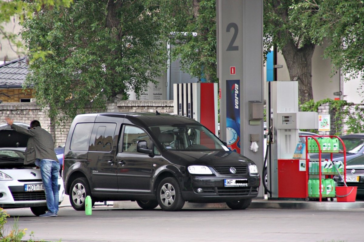 Na stacji paliw widać dwa stojące przy dystrybutorach samochody. Jedno z aut jest ciemne, drugie jasnego koloru. Przy jasnym samochodzi widać mężczyznę, otwierającego klapę silnika.