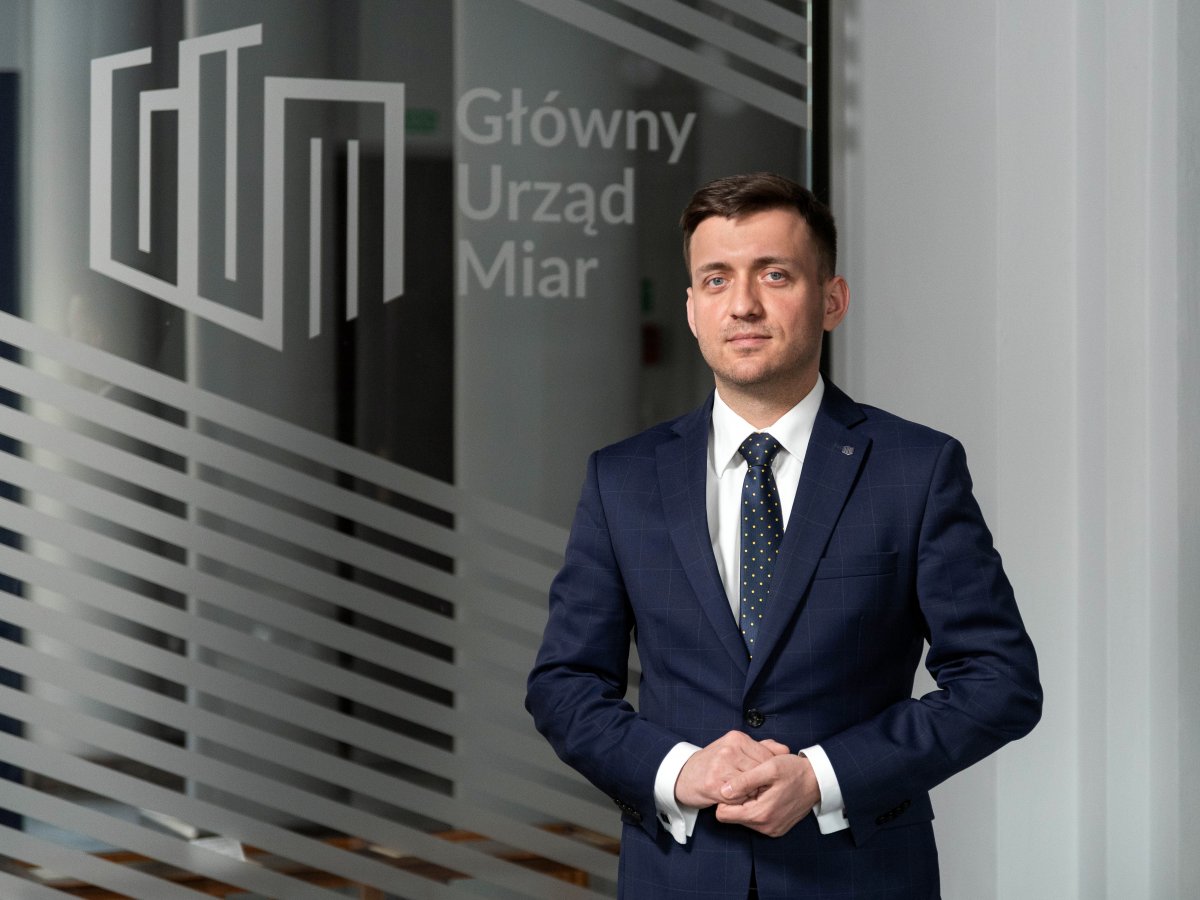 Piotr Ziółkowski - dyrektor generalny głównego urzędu miar od lutego 2021 roku. Młody mężczyzna w garniturze stoi z rękami złożonymi po środku. Z tyłu, za nim, logo GUM na szybie..