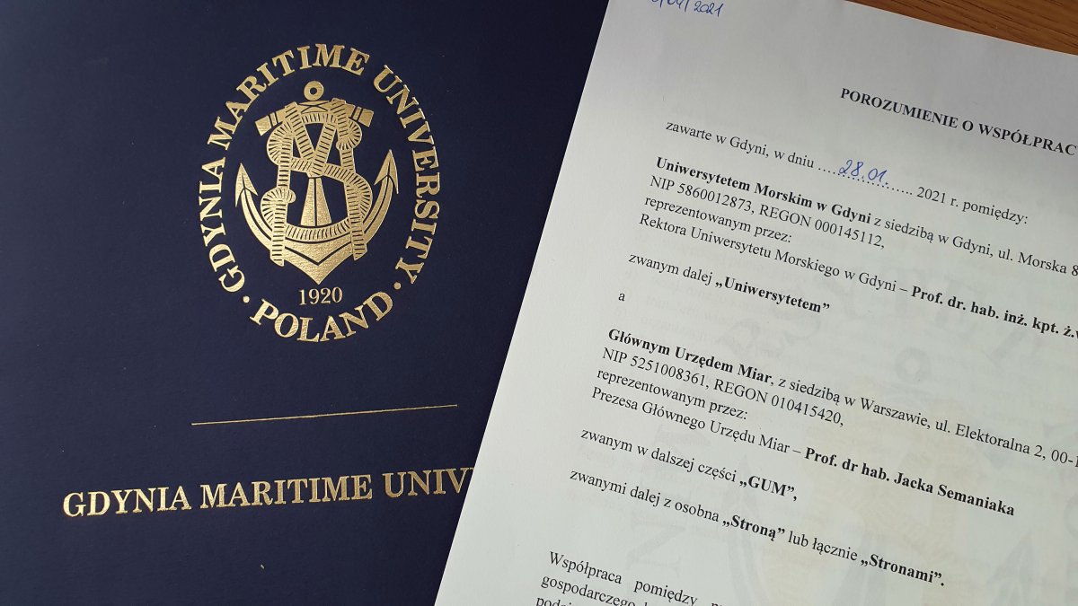 Umowa współpracy między Uniwersytetem Morskim w Gdyni a Głównym Urzędem Miar. Na zdjęciu po prawej stronie kartka z tekstem porozumienia. Po lewej stronie obwoluta teczki firmowej Uniwersytetu Morskiego w Gdyni.