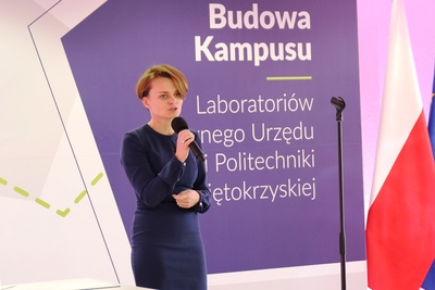  Szczupła kobieta mówi do mikrofonu. Z tyłu, za nią widać tablicę z napisem: Budowa Kampusu Laboratoriów Głównego Urzędu Miar i Politechniki Świętokrzyskiej.