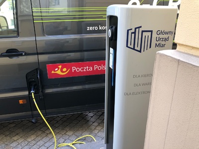  Biała ładowarka do pojazdów elektrycznych podłączona do ciemnego samochodu Poczty Polskiej.