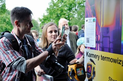  Kobieta i mężczyzna stoją przy plakacie z napisem kamery termowizyjne. Mężczyzna trzyma w ręku aparat fotograficzny oraz telefon.