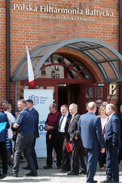  Kilkanaście osób stoi przy ceglastym budynku, na którym widnieje napis Filharmonia Bałtycka.