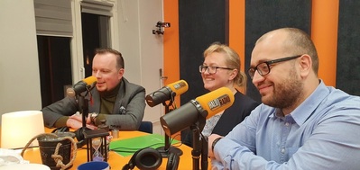  Trzy uśmiechnięte osoby (dwóch mężczyzn i kobieta) siedzą przy mikrofonach w kolorach pomarańczowych. Na mikrofonie widać napis Halo radio.