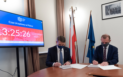  Na zdjęciu dwóch mężczyzn w garniturach i maseczkach siedzi przy stole i podpisuje dokumenty. Z lewej strony widać monitor z wyświetlonym czasem urzędowym, nadzorowanym w GUM. Za mężczyznami flagi Polski i Unii Europejskiej, na ścianie stare zdjęcie.