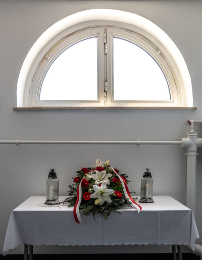  Zdjęcie okna do wolności - Pod oknem, na stoliku leży wieniec kwiatów przewiązany biało-czerwoną szarfą, po bokach stoją zapalone znicze.