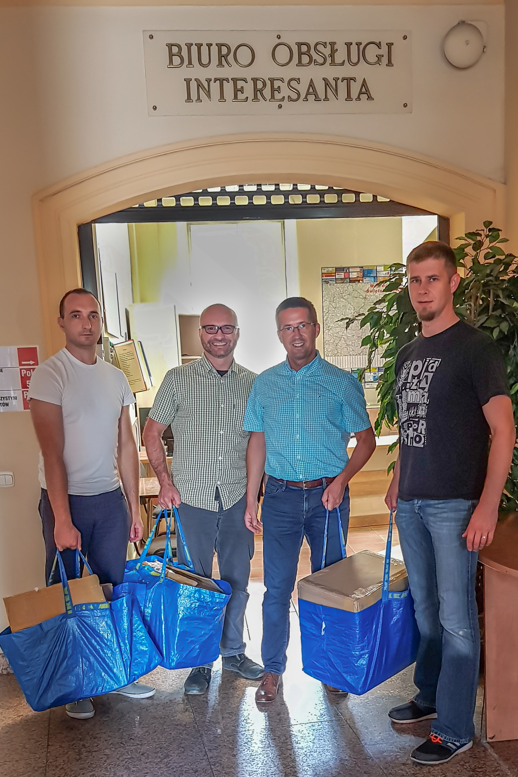 4 mężczyzn stoi z niebieskimi torbami wypełnionymi pudłami. Nad nimi napis Biuro Obsługi Interesanta.